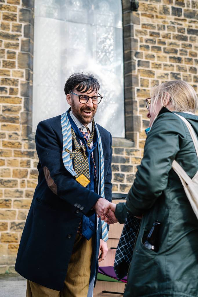 Charlie, a Caucasian man with dark hair and a short beard, is shaking the hand of an audience member. They are both outside in fron of a church. Charlie is wearing beige trousers, a long navy winters coat, a shirt and several ties.