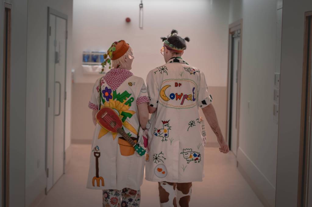 A hospital corridor with two adults walking away from the camera. Both figures wear white doctors coats with colourful writing and images. On the left - Dr Flowerpot and on the right - Charlie as Dr Cowpat