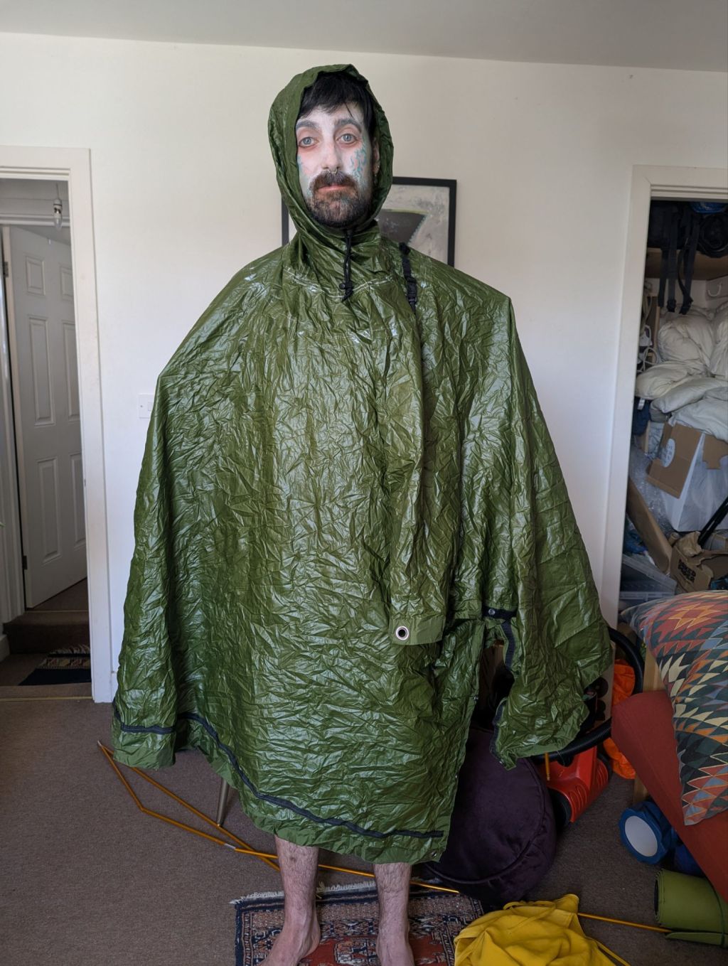 A man dressed in a green poncho stands looking a the camera. He has white makeup on his face and red around the eyes. He is standing in a living room.