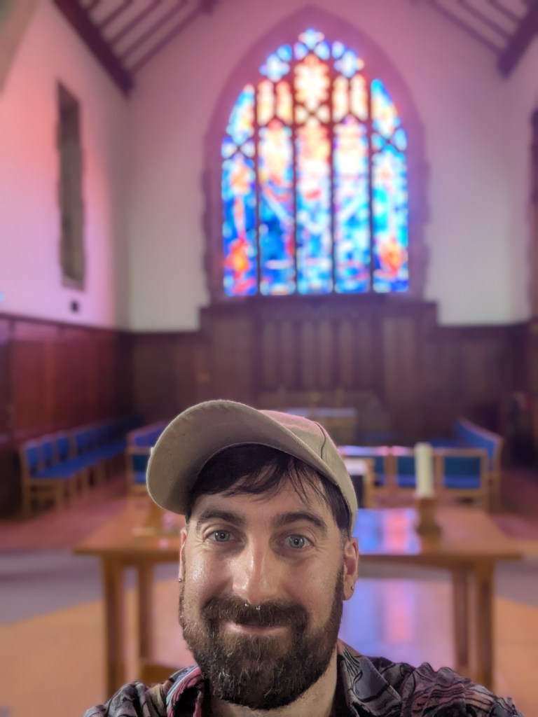 Charlie inside a church, wearing a pale cap, smiling at the camera with a stained glass windows behind him.