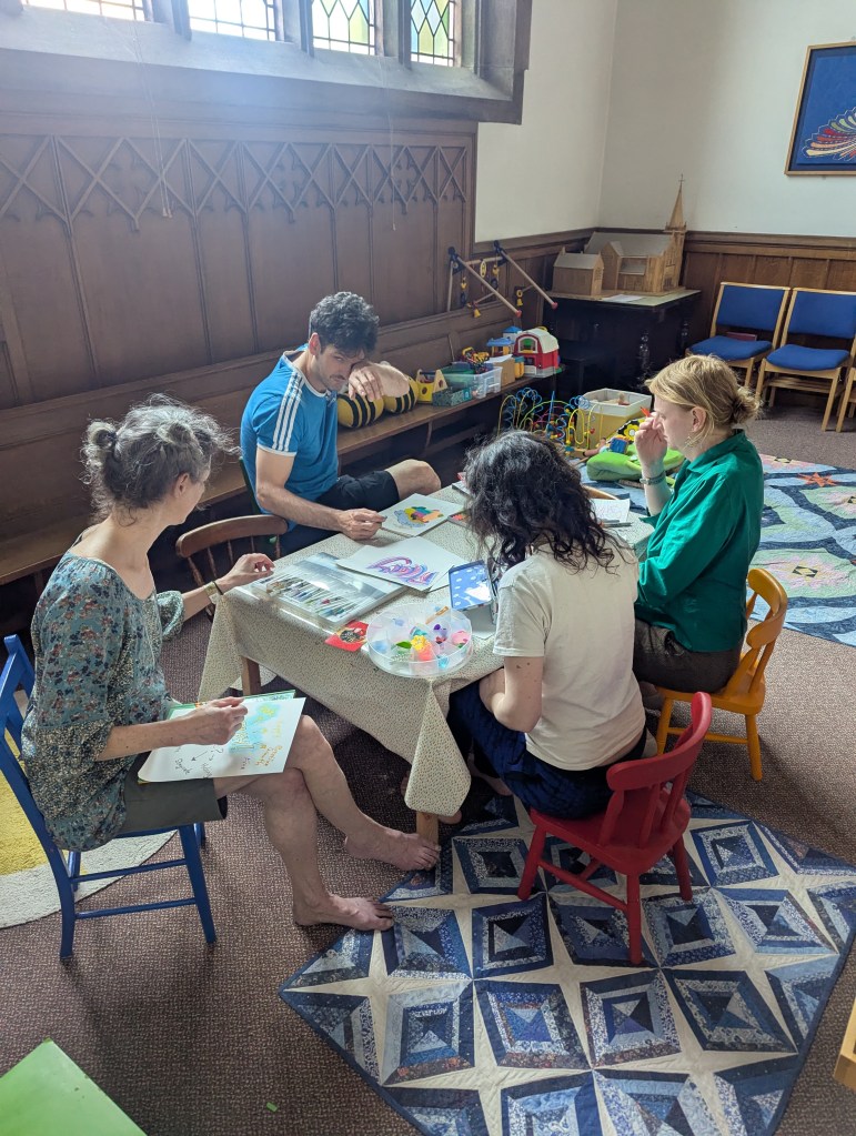 4 adults sit around a children's table on coloured chairs making artwork.
