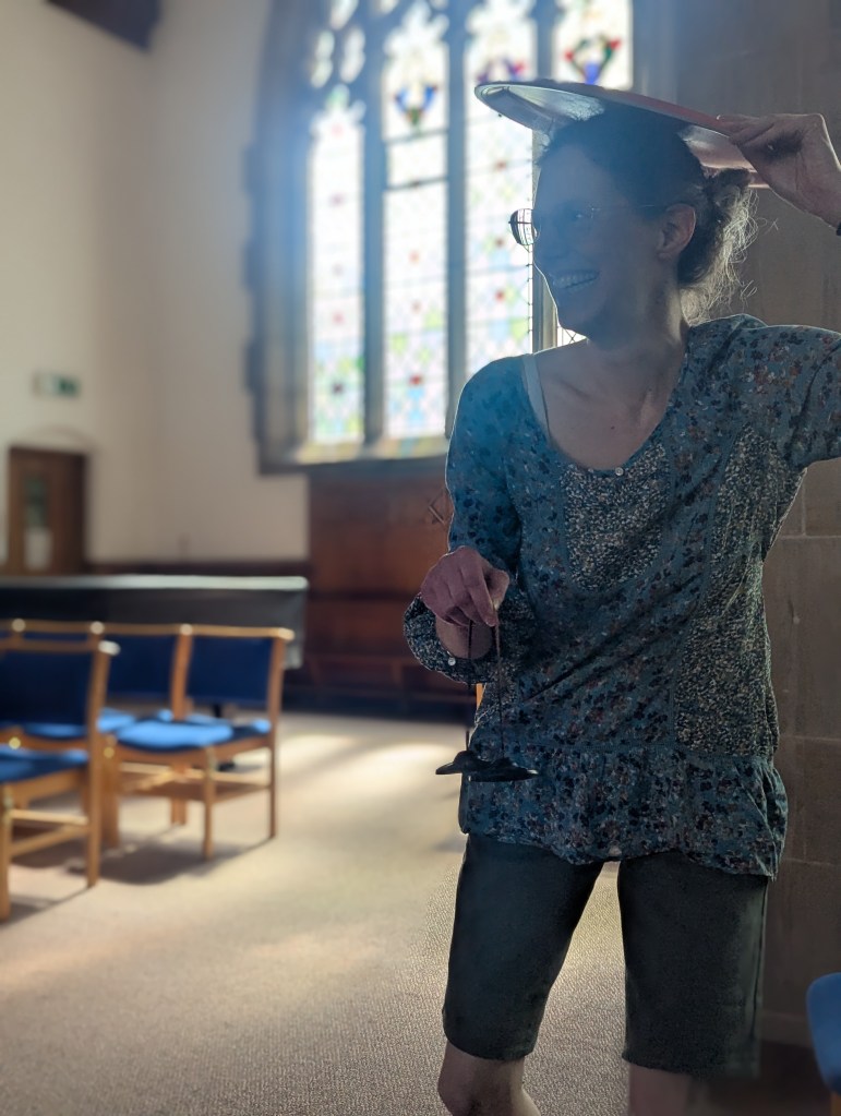 A woman stands near a church window, a set of bells in one hand and a round wooden tray held on her head in the other hand.