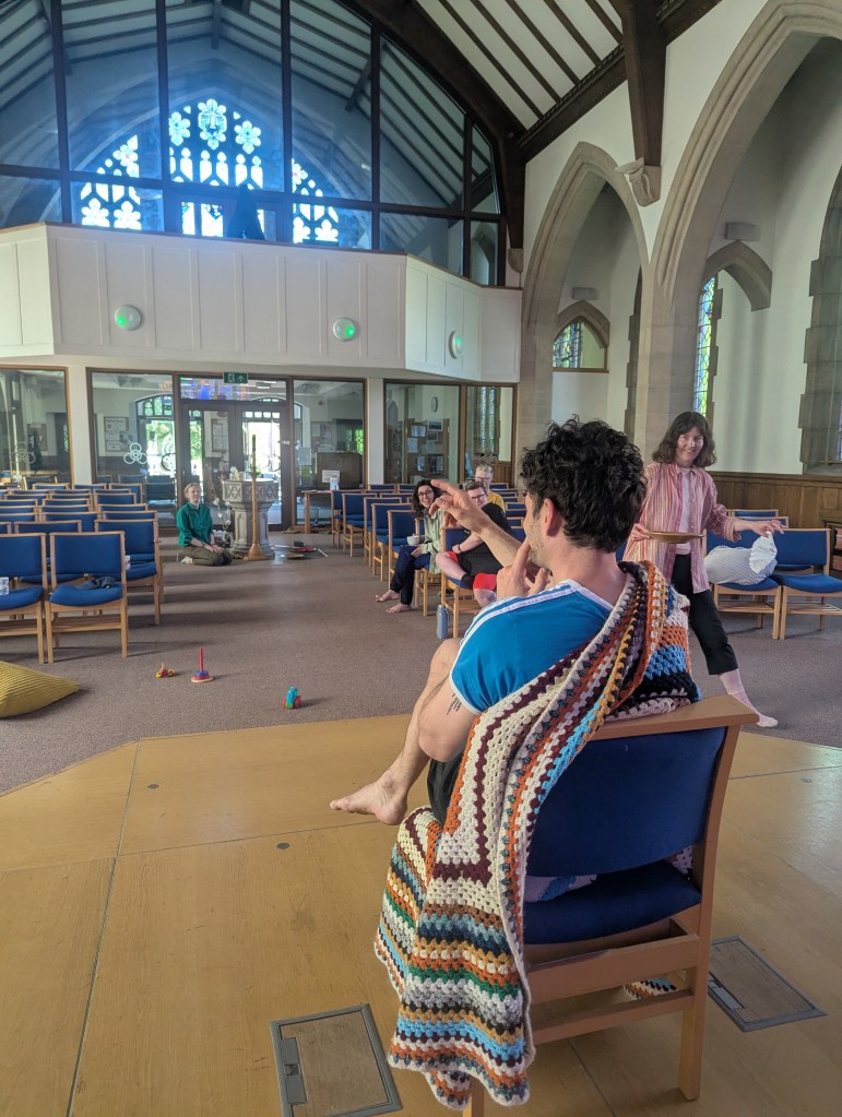 Inside a church, one participant sits eye closed on a chair pointing to empty air, whilst another participant is seen stepping to the right holding two objects. Several other objects are scattered on the floor. The other participants sit on chairs watching the game and laughing.
